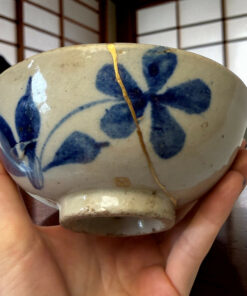 Close-up of a blue-and-white ceramic plate with a gold fault-line crack repaired in the center, held by hands.