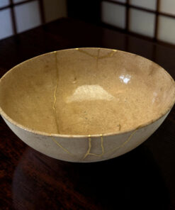 Close-up of a beige speckled ceramic bowl with a delicate gold line design, placed on a dark wooden table with blurred room panels in the background