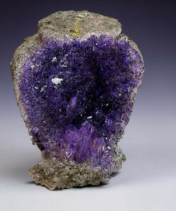 A purple amethyst mineral cluster with crystalline facets, set against a muted gray gradient background.