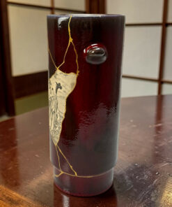 Close-up of a dark red lacquered cylinder with a gold crackle seam and torn label, resting on a wooden table.