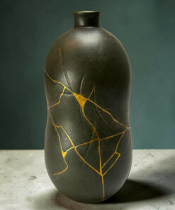 Dark matte ceramic vase with gold repair lines (kintsugi) running through the cracks, set on a light speckled floor against a muted backdrop.