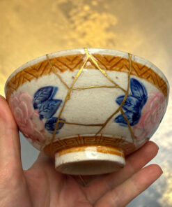 Close-up of a cracked ceramic object repaired with gold lacquer (kintsugi) being held in a hand, with a golden blurred background.