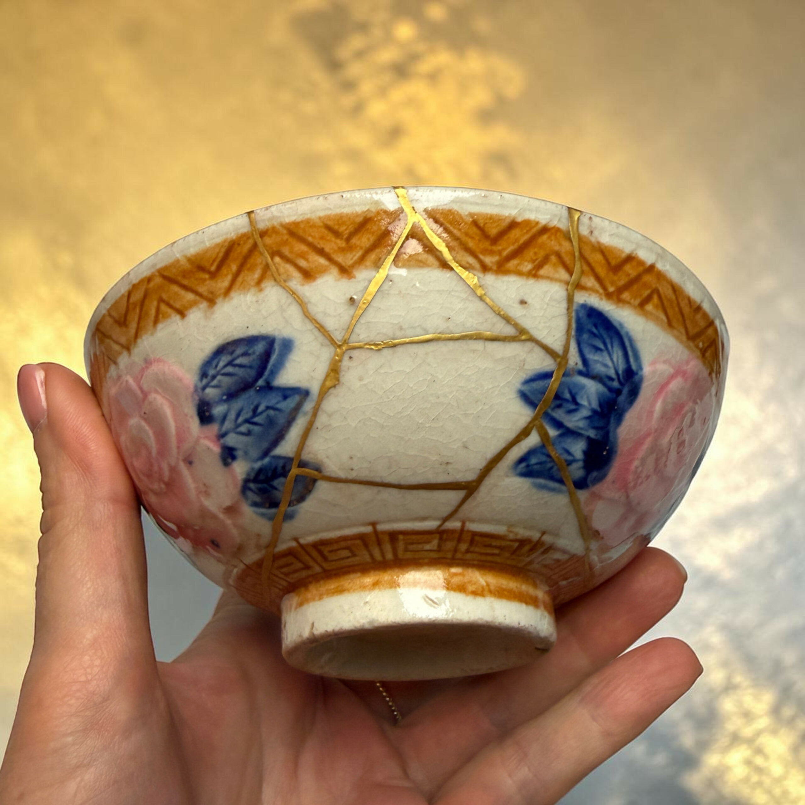 IMG_2496-Photoroom | Kintsugi by Myriam GREFF Close-up of a cracked ceramic object repaired with gold lacquer (kintsugi) being held in a hand, with a golden blurred background.