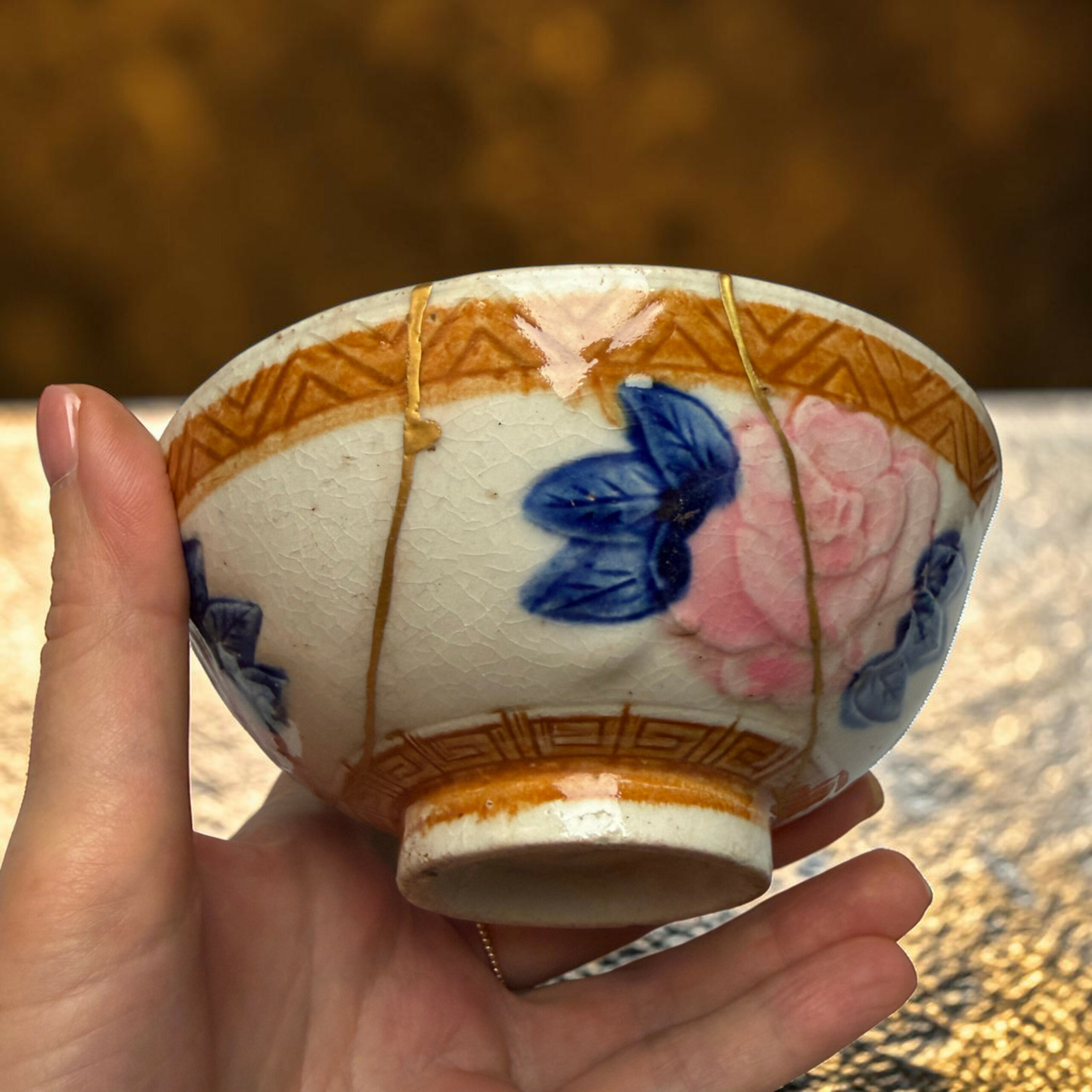 IMG_2497-Photoroom | Kintsugi by Myriam GREFF Close-up of a hand holding a decorated ceramic egg with blue and pink floral patterns and gold lines.