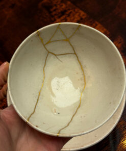 Close-up of a light ceramic bowl with gold repair seams along the rim, held by a hand against a dark wooden background.