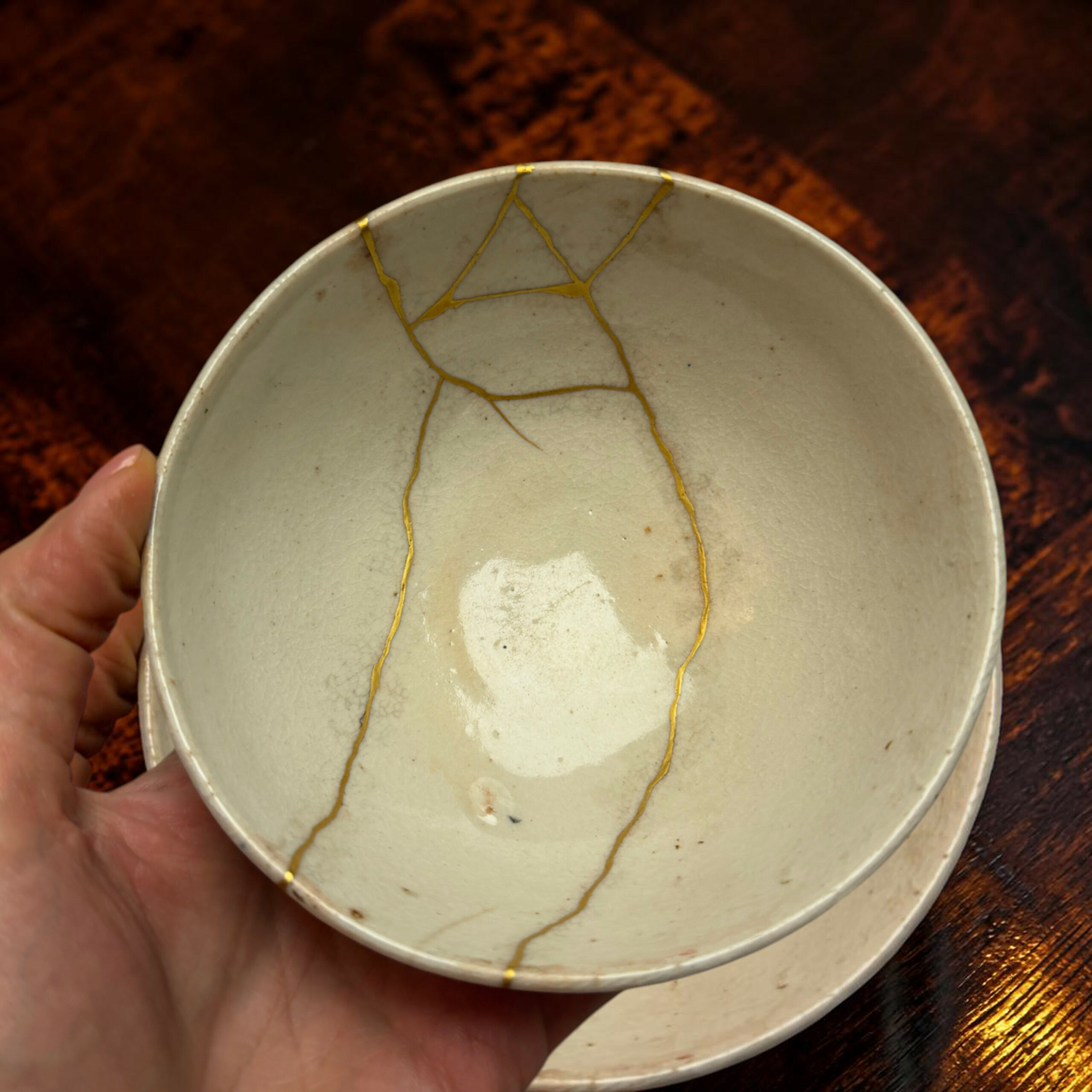 IMG_2498-Photoroom | Kintsugi by Myriam GREFF Close-up of a light ceramic bowl with gold repair seams along the rim, held by a hand against a dark wooden background.