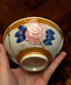 Hand holding a decorative porcelain egg with pink and blue floral patterns and a gold string against a dark wooden surface.