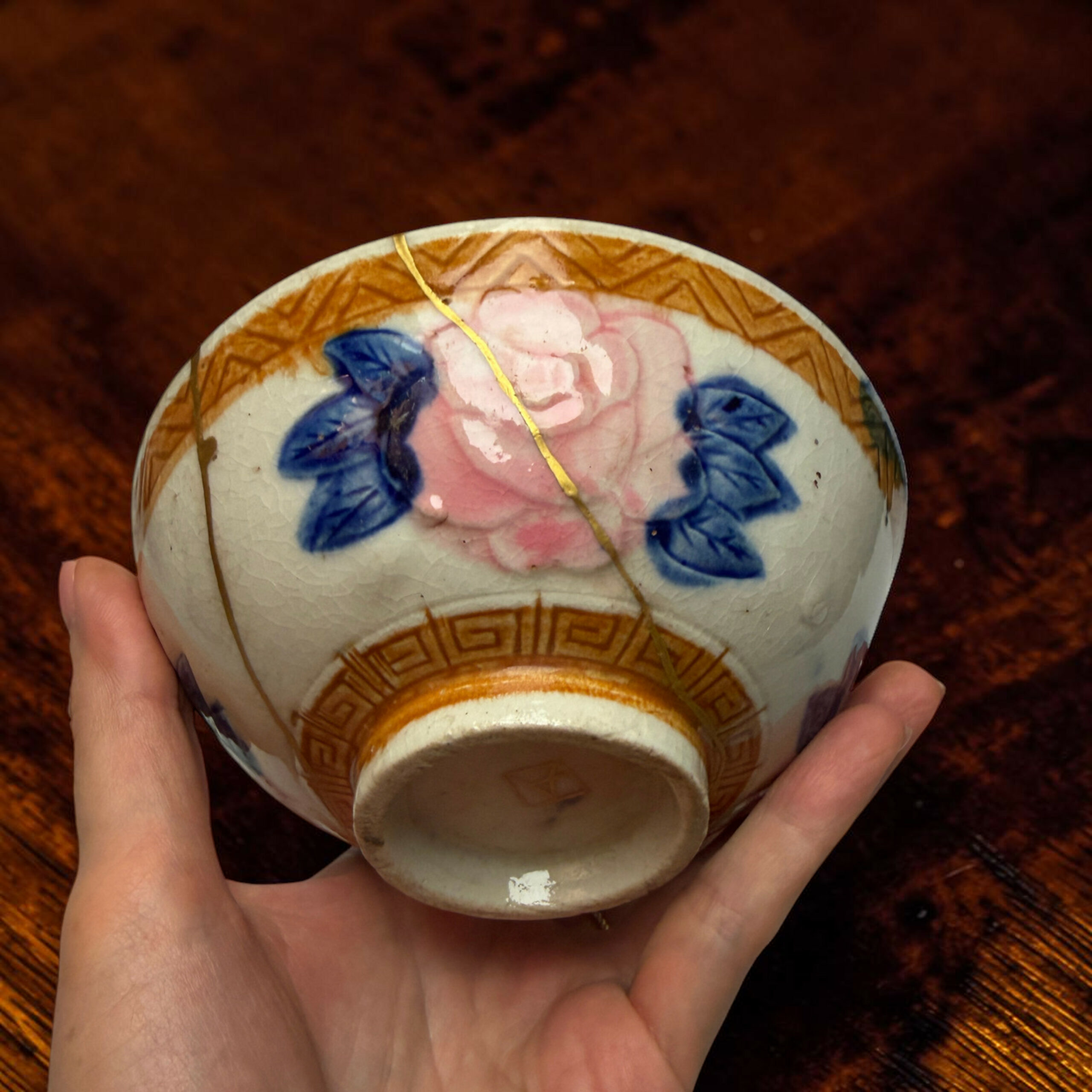 IMG_2499-Photoroom | Kintsugi by Myriam GREFF Hand holding a decorative porcelain egg with pink and blue floral patterns and a gold string against a dark wooden surface.