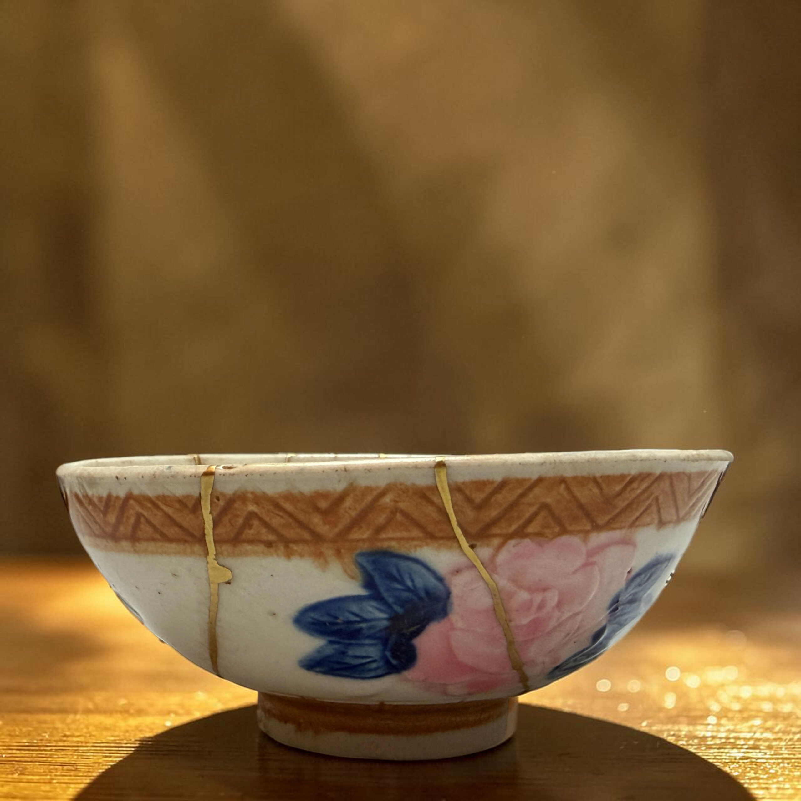 IMG_2501-Photoroom | Kintsugi by Myriam GREFF Decorative ceramic bowl with blue and pink floral pattern, hung by cords above a wooden table in warm light.