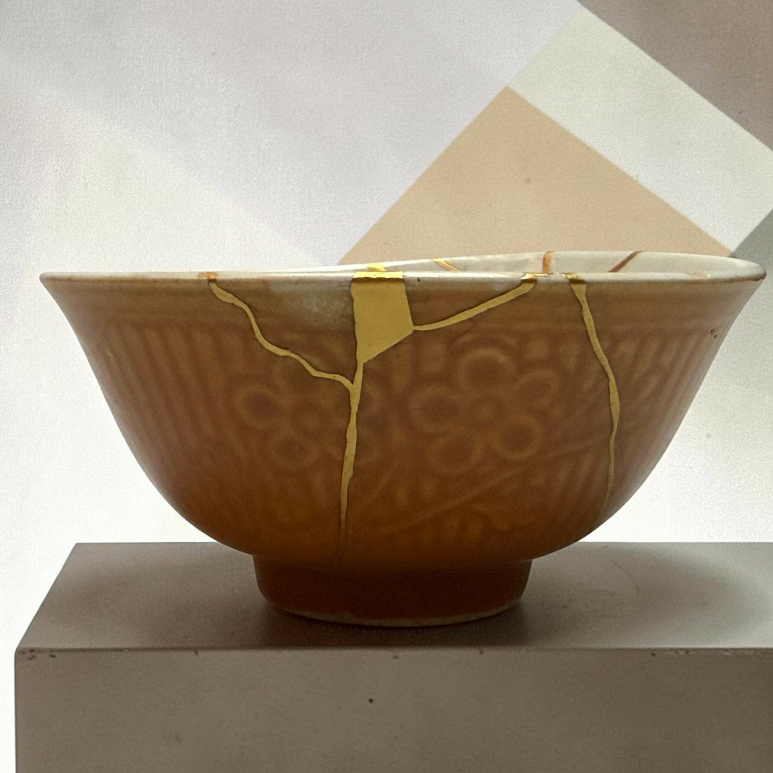 IMG_2502-Photoroom | Kintsugi by Myriam GREFF Close-up of a brown ceramic planter with yellow straps, on a light pedestal against a pale geometric background.