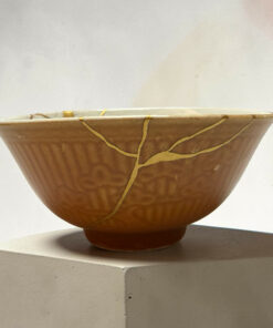 Close-up of a brown ceramic bowl with gold-kintsugi cracks along the rim, resting on a concrete pedestal against a light background