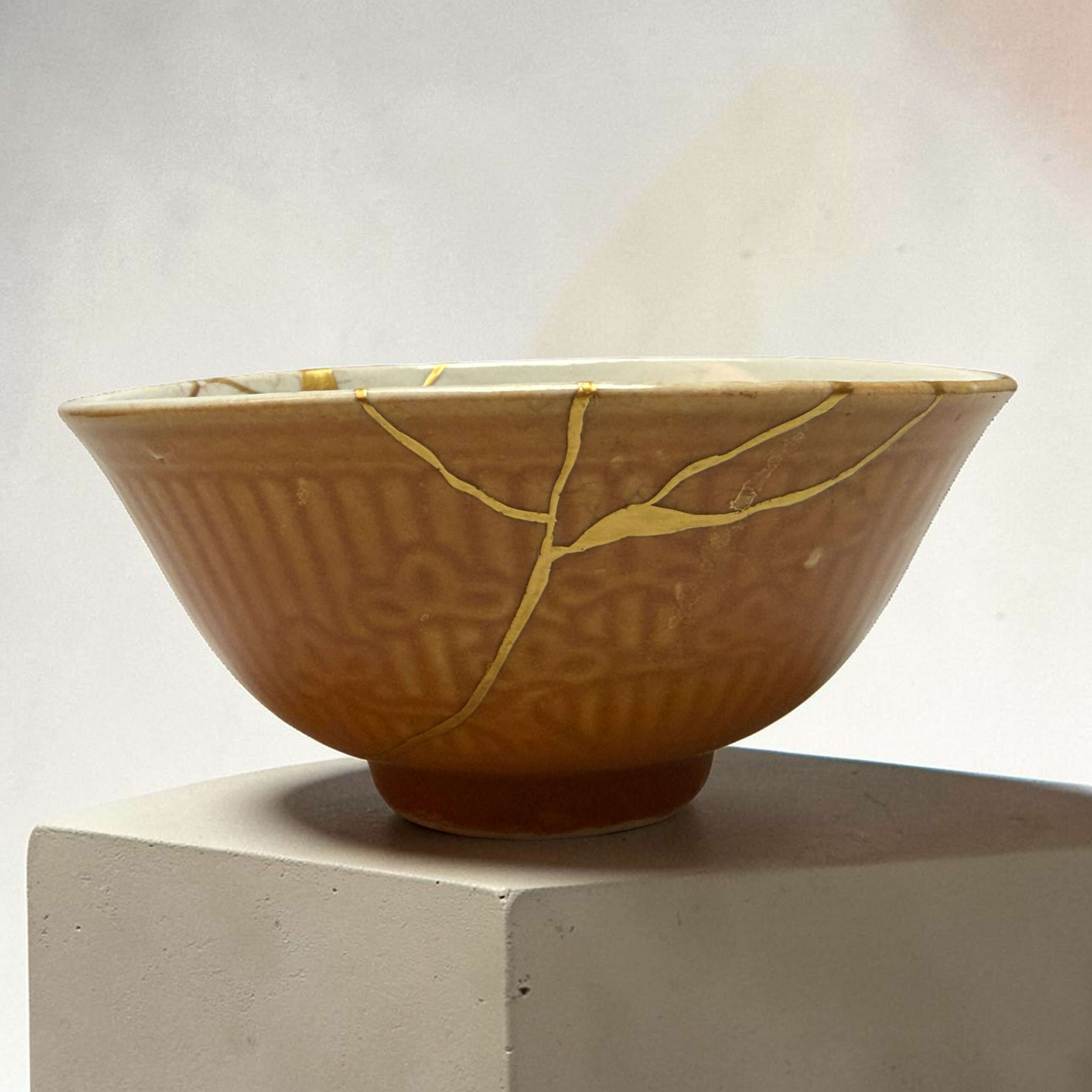 IMG_2504-Photoroom | Kintsugi by Myriam GREFF Close-up of a brown ceramic bowl with gold-kintsugi cracks along the rim, resting on a concrete pedestal against a light background