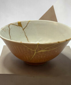 Close-up of a beige ceramic bowl with gold string wrapping, resting on a white block.