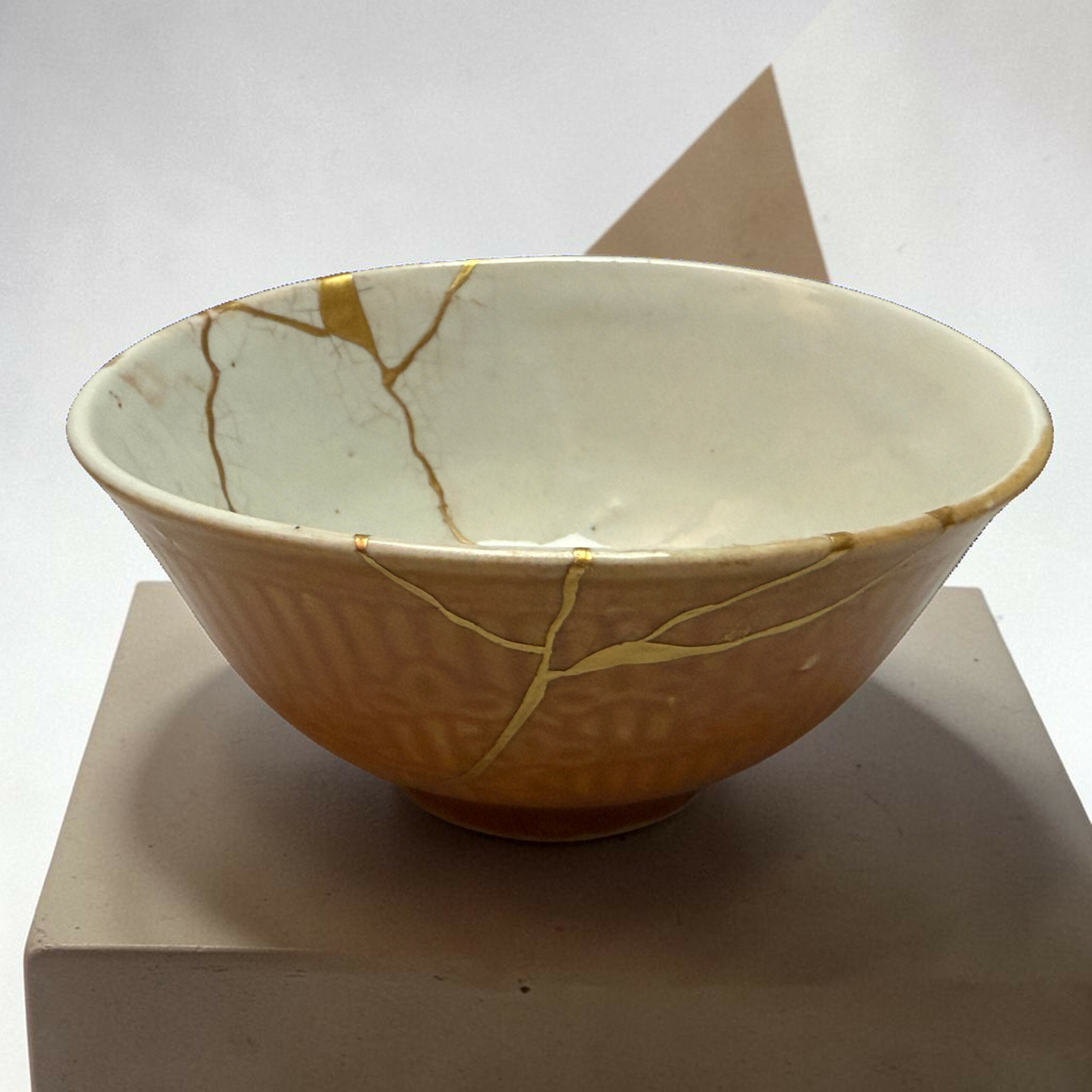 IMG_2505-Photoroom | Kintsugi by Myriam GREFF Close-up of a beige ceramic bowl with gold string wrapping, resting on a white block.