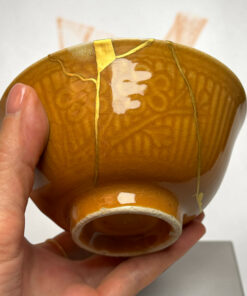 Close-up of a small, cracked brown ceramic object held in a hand, with yellow glaze lines and a curved edge visible.