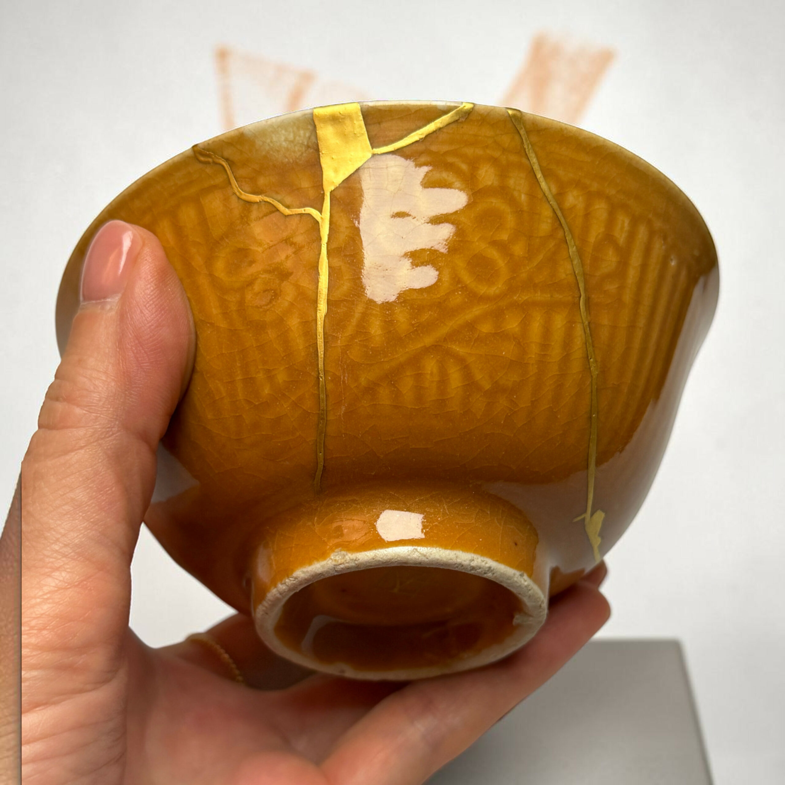 IMG_2507-Photoroom | Kintsugi by Myriam GREFF Close-up of a small, cracked brown ceramic object held in a hand, with yellow glaze lines and a curved edge visible.