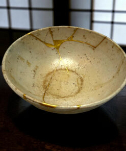 Close-up of a white ceramic bowl with gold kintsugi cracks on a dark wooden table, window grid blurred in background.