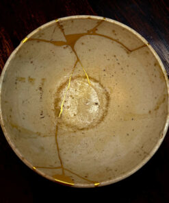 Round ceramic plate with gold-kintsugi repairs, where gold seams highlight cracks on a light glaze, resting on a dark wooden surface.
