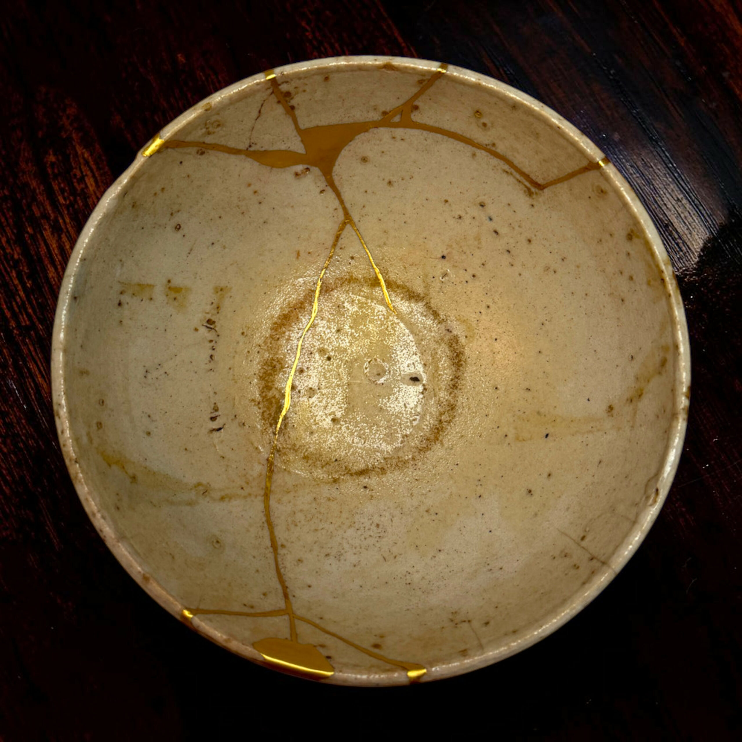IMG_2509-Photoroom | Kintsugi by Myriam GREFF Round ceramic plate with gold-kintsugi repairs, where gold seams highlight cracks on a light glaze, resting on a dark wooden surface.