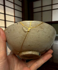 Close-up of a person’s hands holding a round, light-gray stone with gold crack lines (kintsugi) against a grid-like background.