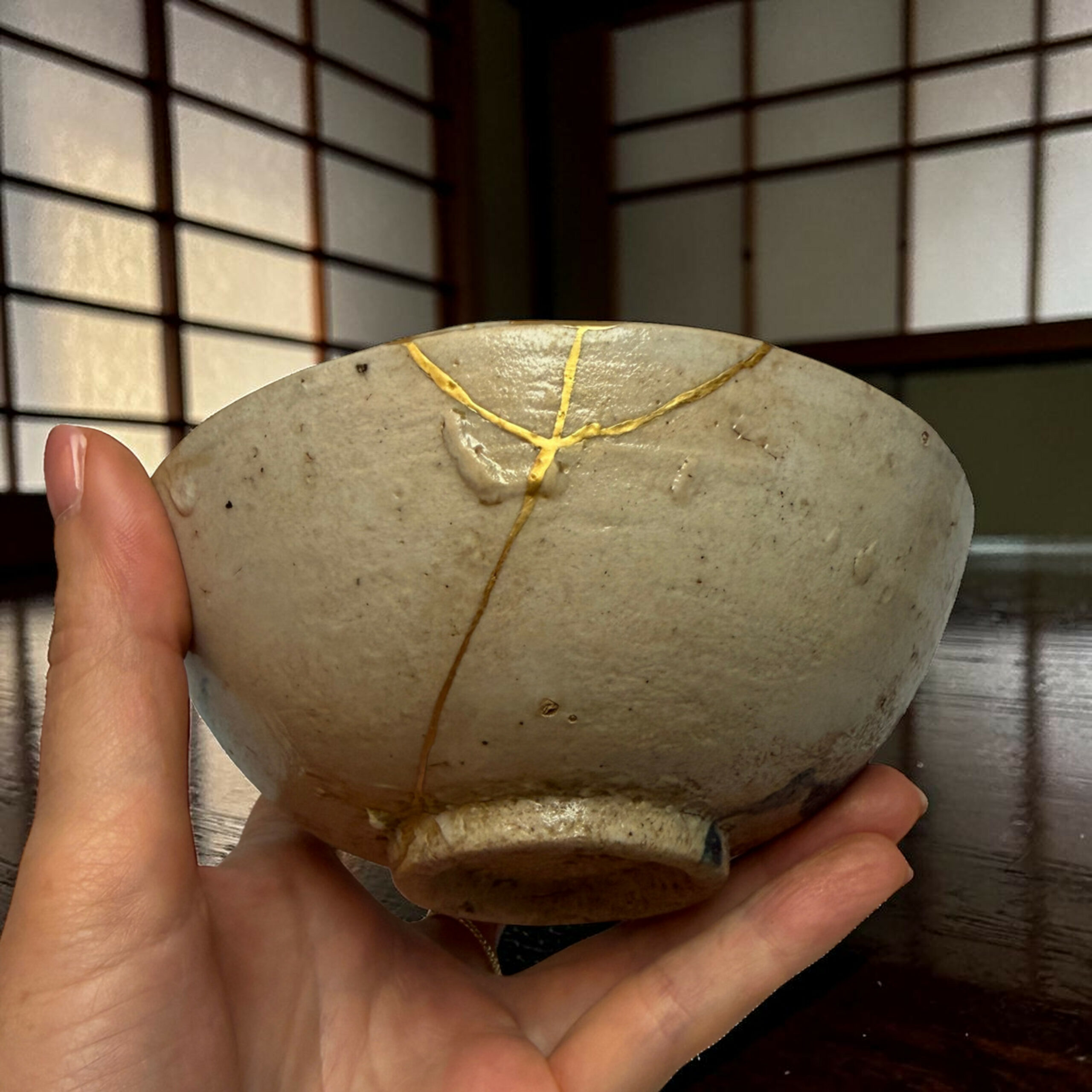 IMG_2510-Photoroom | Kintsugi by Myriam GREFF Close-up of a person’s hands holding a round, light-gray stone with gold crack lines (kintsugi) against a grid-like background.