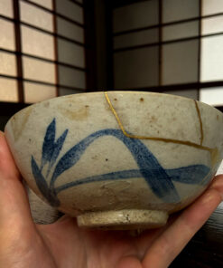 A hand holds a round, light-colored ceramic bowl with blue abstract designs and a thin yellow string looping across its surface; dark grid-patterned background behind.
