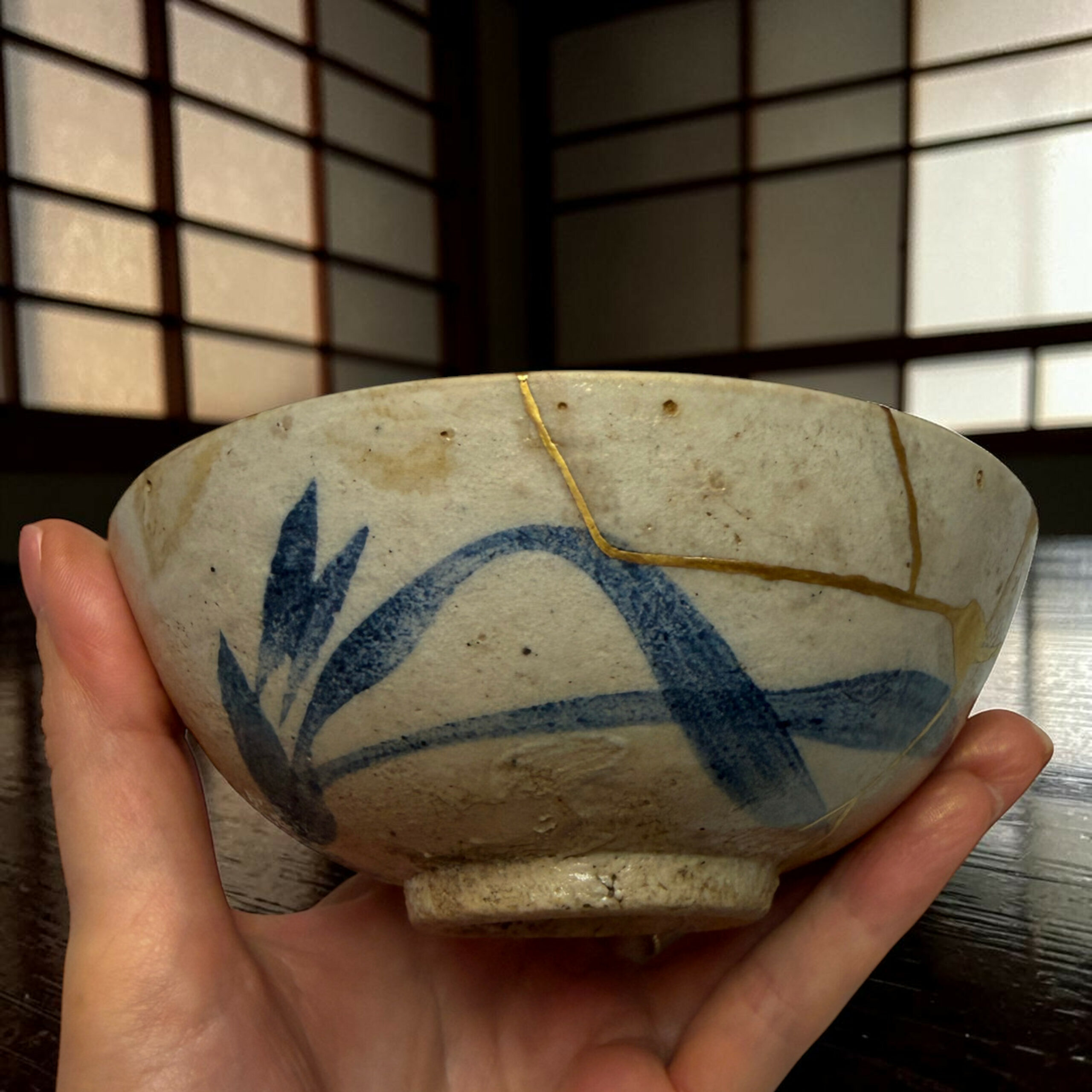 IMG_2511-Photoroom | Kintsugi by Myriam GREFF A hand holds a round, light-colored ceramic bowl with blue abstract designs and a thin yellow string looping across its surface; dark grid-patterned background behind.