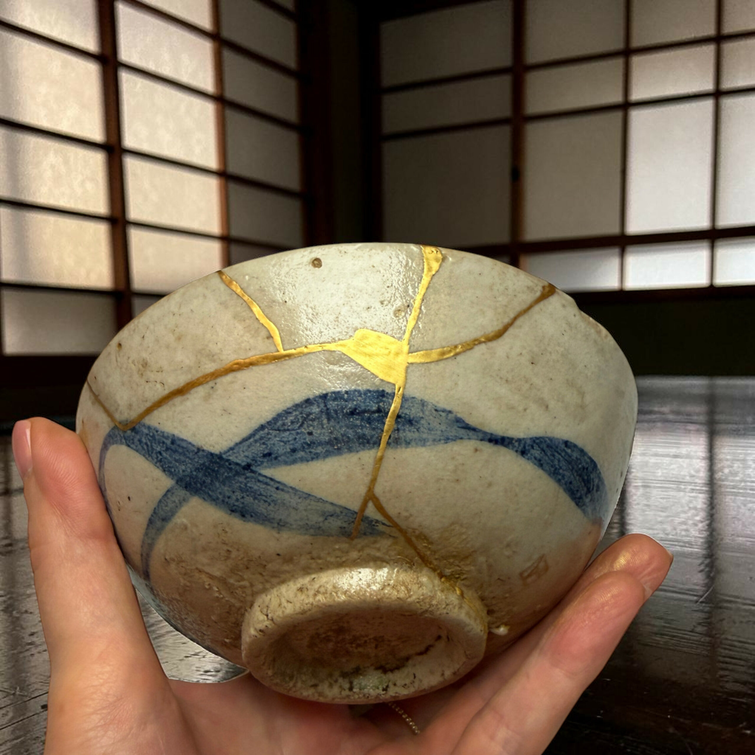 IMG_2512-Photoroom | Kintsugi by Myriam GREFF Close-up of a hand holding a smooth stone with gold-veined cracks, set against a dark, slatted background.