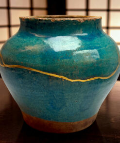 Close-up of a blue ceramic vase with a crackled glaze and a gold rope motif, against a dark, grid-patterned background.