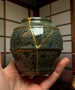 Close-up of a mottled, crackled ceramic vase with gold ribbon seams, held in a person’s hands indoors.