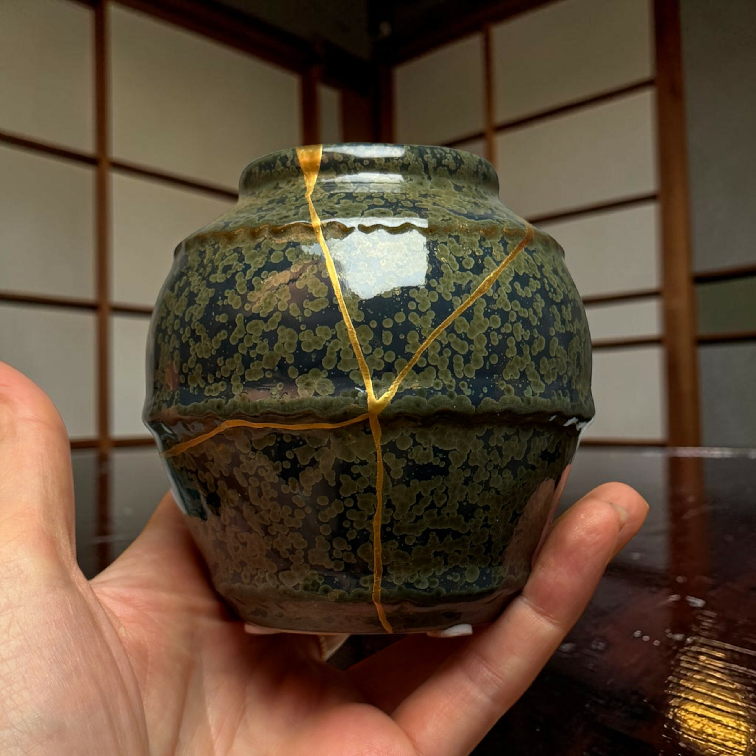 IMG_2524-Photoroom | Kintsugi by Myriam GREFF Close-up of a mottled, crackled ceramic vase with gold ribbon seams, held in a person’s hands indoors.