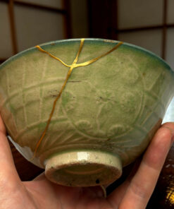 Close-up of a green ceramic bowl repaired with gold seams (kintsugi), held in a person's hands.