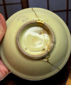 Close-up of a pale green ceramic plate with a long crack repaired with gold kintsugi, held by a person's fingers at the edge.