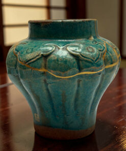 Close-up of a turquoise ceramic vase with carved patterns and gold lines, sitting on a dark wooden table