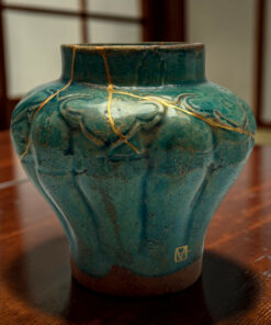 Close-up of a teal ceramic vase with gold crackle lines and a dark brown base on a wooden table (kintsugi-inspired repair).