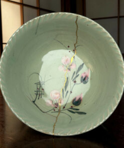 Close-up of a pale green ceramic plate with a gold crack running down the rim, floral pattern, on a wooden table.