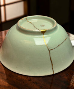 Green ceramic vase with gold kintsugi cracks resting on a wooden surface, close-up detail of repaired pottery.