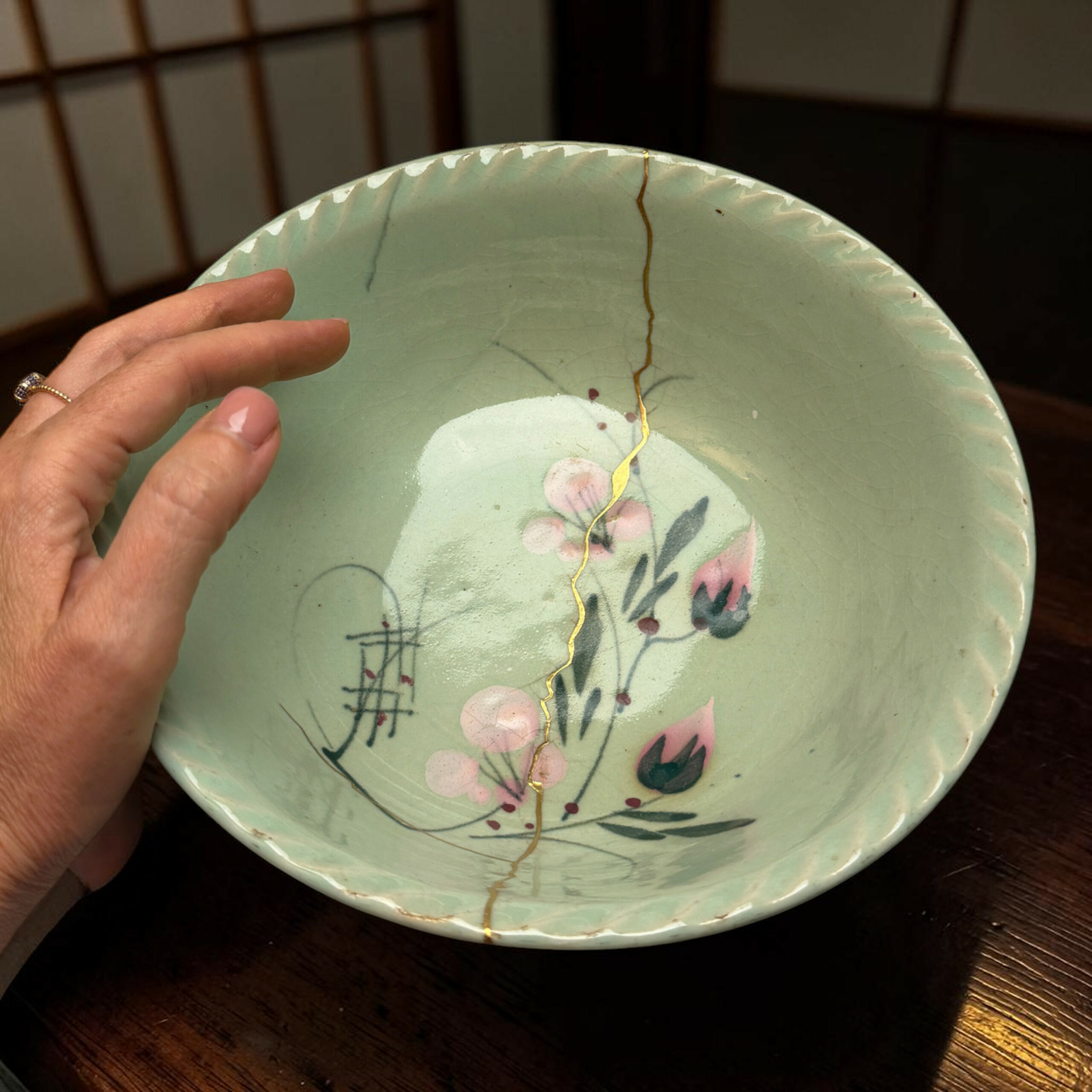 IMG_2542-Photoroom | Kintsugi by Myriam GREFF Close-up of a pale green ceramic plate with floral design, showing a long crack and a hand resting on its edge.