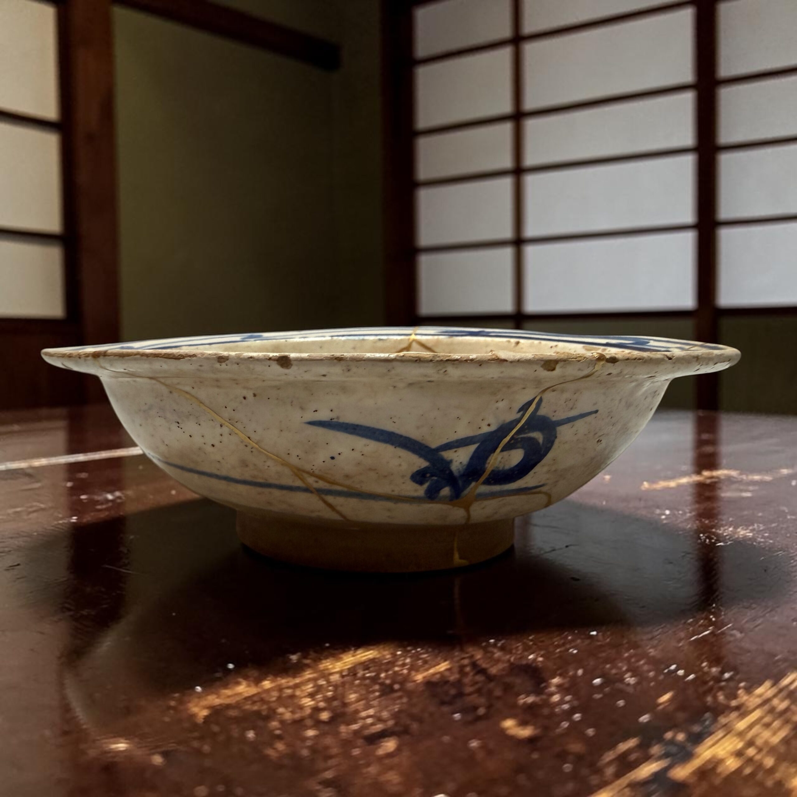 IMG_2551 | Kintsugi by Myriam GREFF Close-up of a blue-and-white ceramic bowl on a polished wooden table with scattered crumbs around it.
