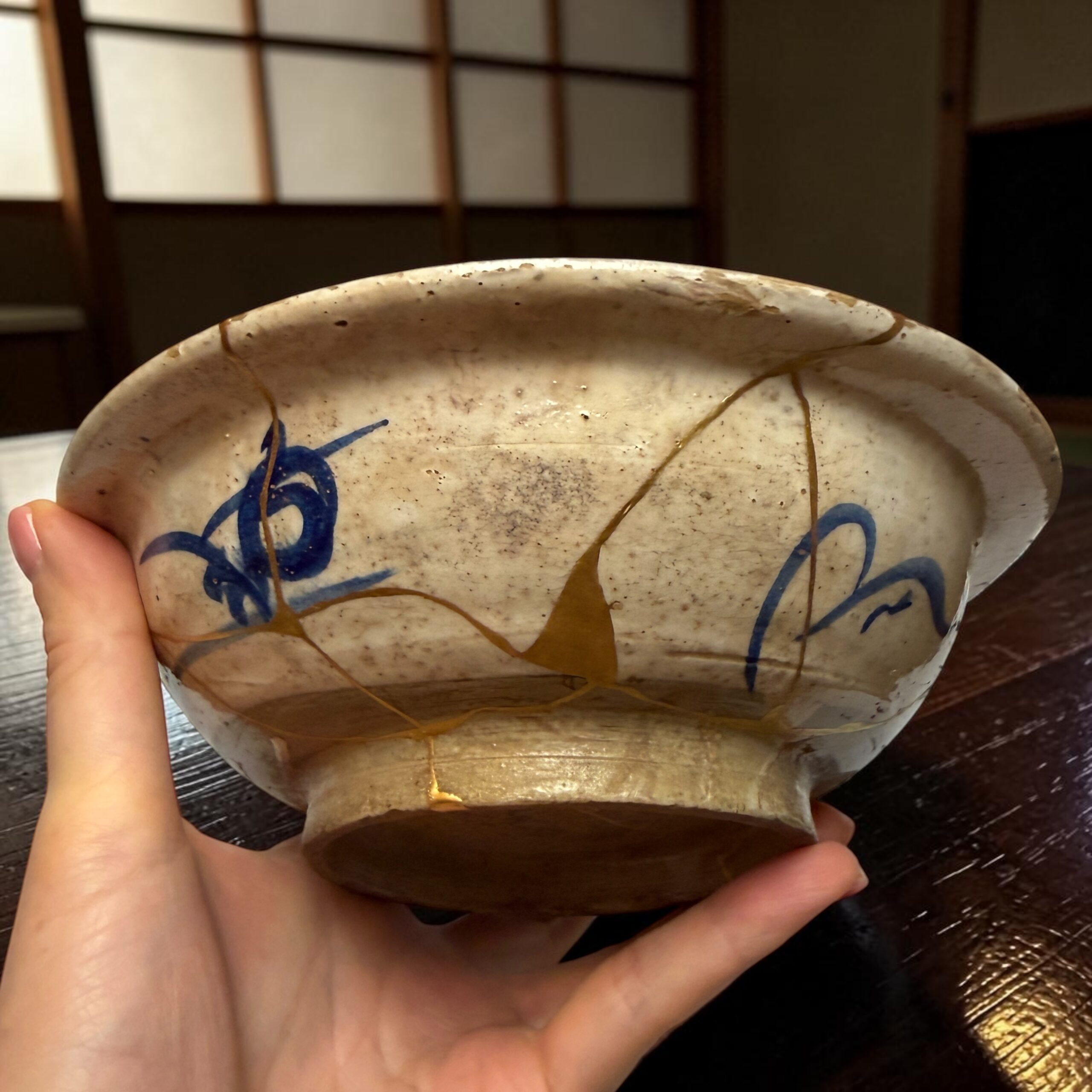 IMG_2553 | Kintsugi by Myriam GREFF Close-up of a speckled ceramic bowl with blue designs being held by a person, indoors at a wooden table.