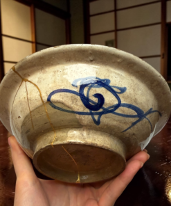 Hands cradle a weathered ceramic bowl with blue glaze and visible crackle lines in a warm setting.