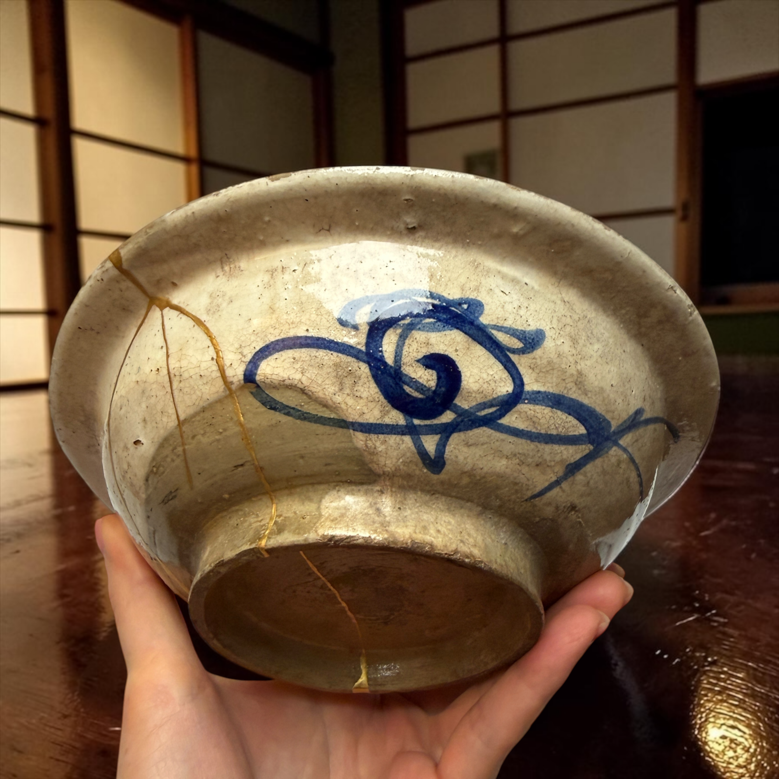 IMG_2554 | Kintsugi by Myriam GREFF Hands cradle a weathered ceramic bowl with blue glaze and visible crackle lines in a warm setting.