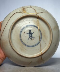 Close-up of a white porcelain plate with blue glaze design, repaired with gold kintsugi cracks, held by a hand.