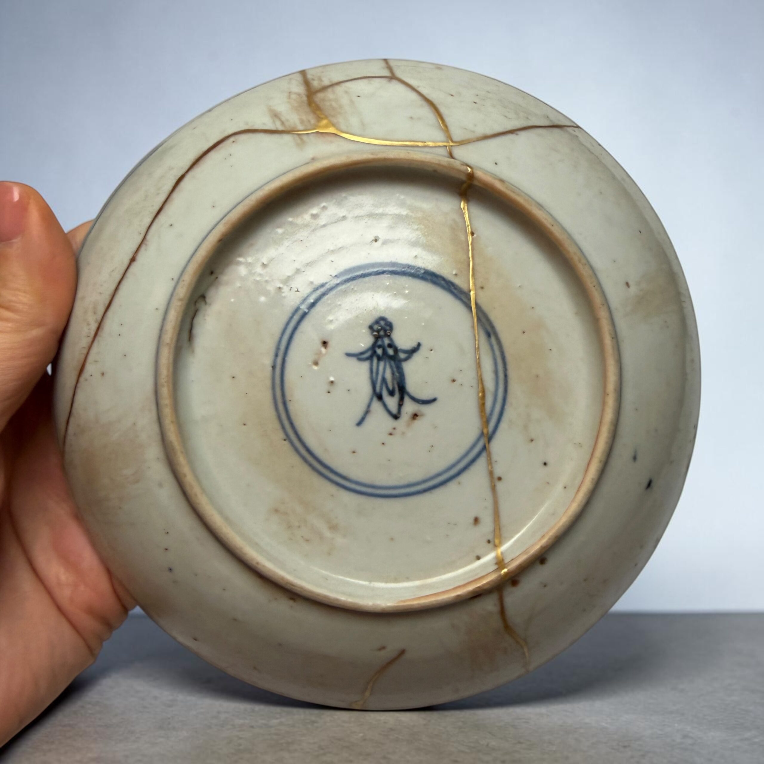 IMG_2557 | Kintsugi by Myriam GREFF Close-up of a white porcelain plate with blue glaze design, repaired with gold kintsugi cracks, held by a hand.