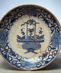 Close-up of a blue-and-white ceramic plate repaired with gold lacquer (kintsugi), showing gold seams along the cracked rim and intricate blue floral patterns.