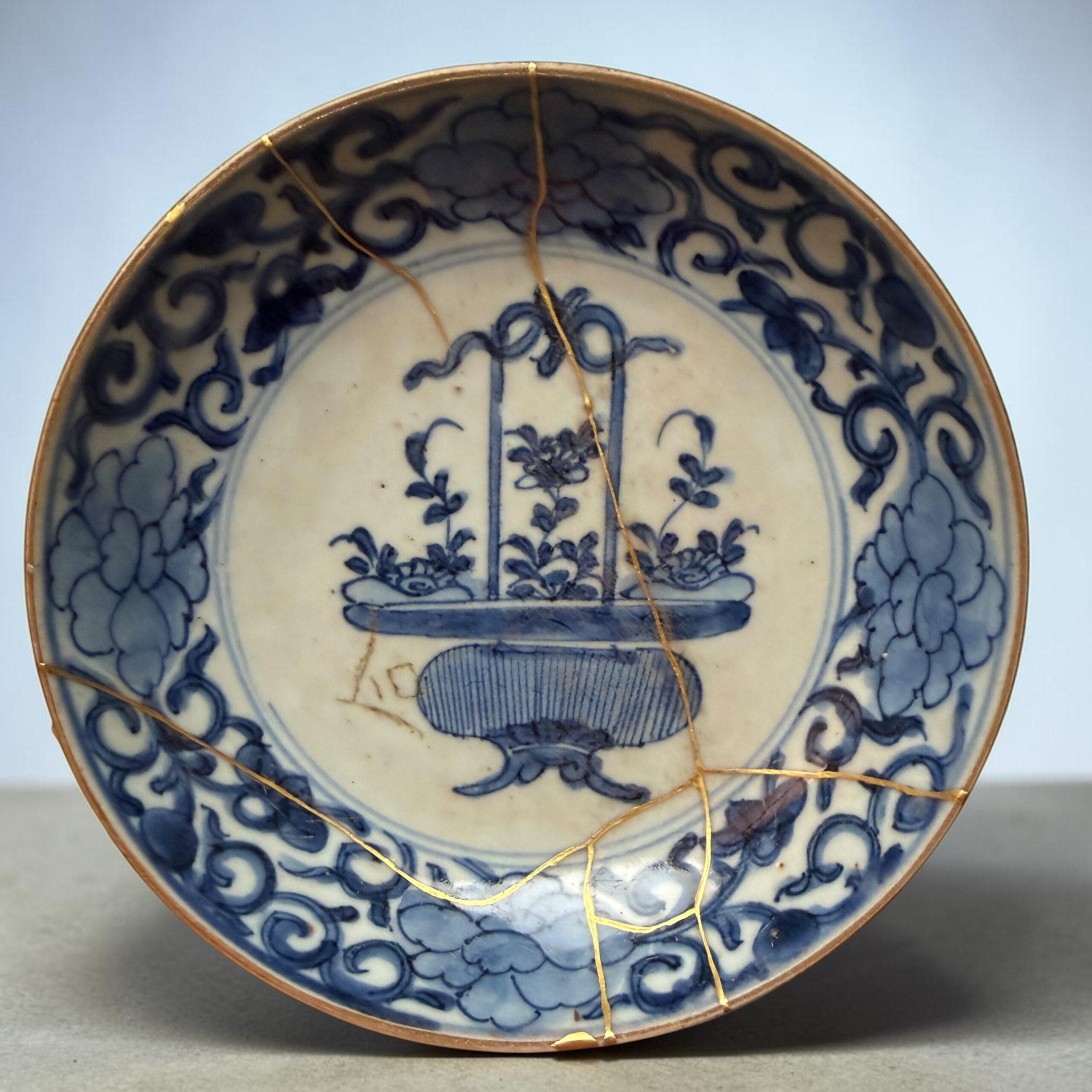 IMG_2558 | Kintsugi by Myriam GREFF Close-up of a blue-and-white ceramic plate repaired with gold lacquer (kintsugi), showing gold seams along the cracked rim and intricate blue floral patterns.