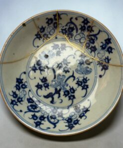 Close-up of a blue-and-white porcelain plate with gold seams, showing repaired cracks (kintsugi) on a pale background.