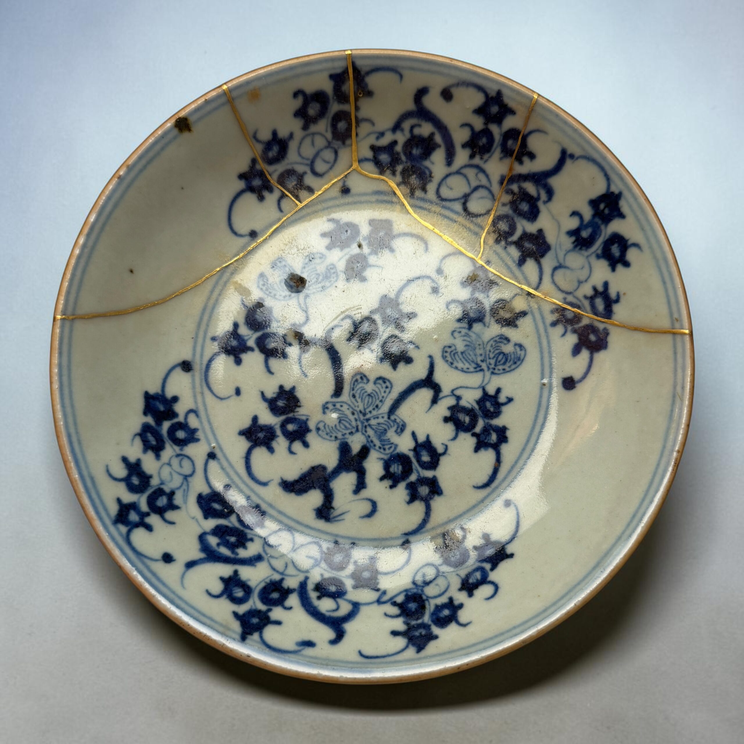 IMG_2559 | Kintsugi by Myriam GREFF Close-up of a blue-and-white porcelain plate with gold seams, showing repaired cracks (kintsugi) on a pale background.