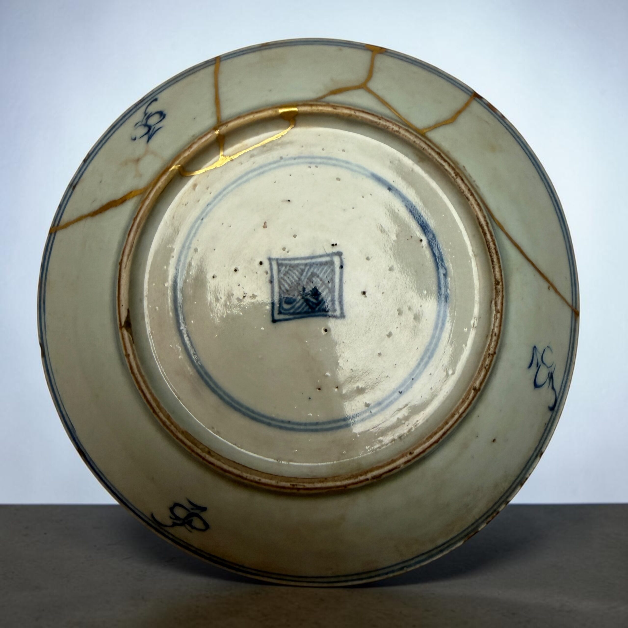 IMG_2560 | Kintsugi by Myriam GREFF Close-up of a ceramic plate repaired with gold seams (kintsugi), showing blue rim and cracks in the glaze.