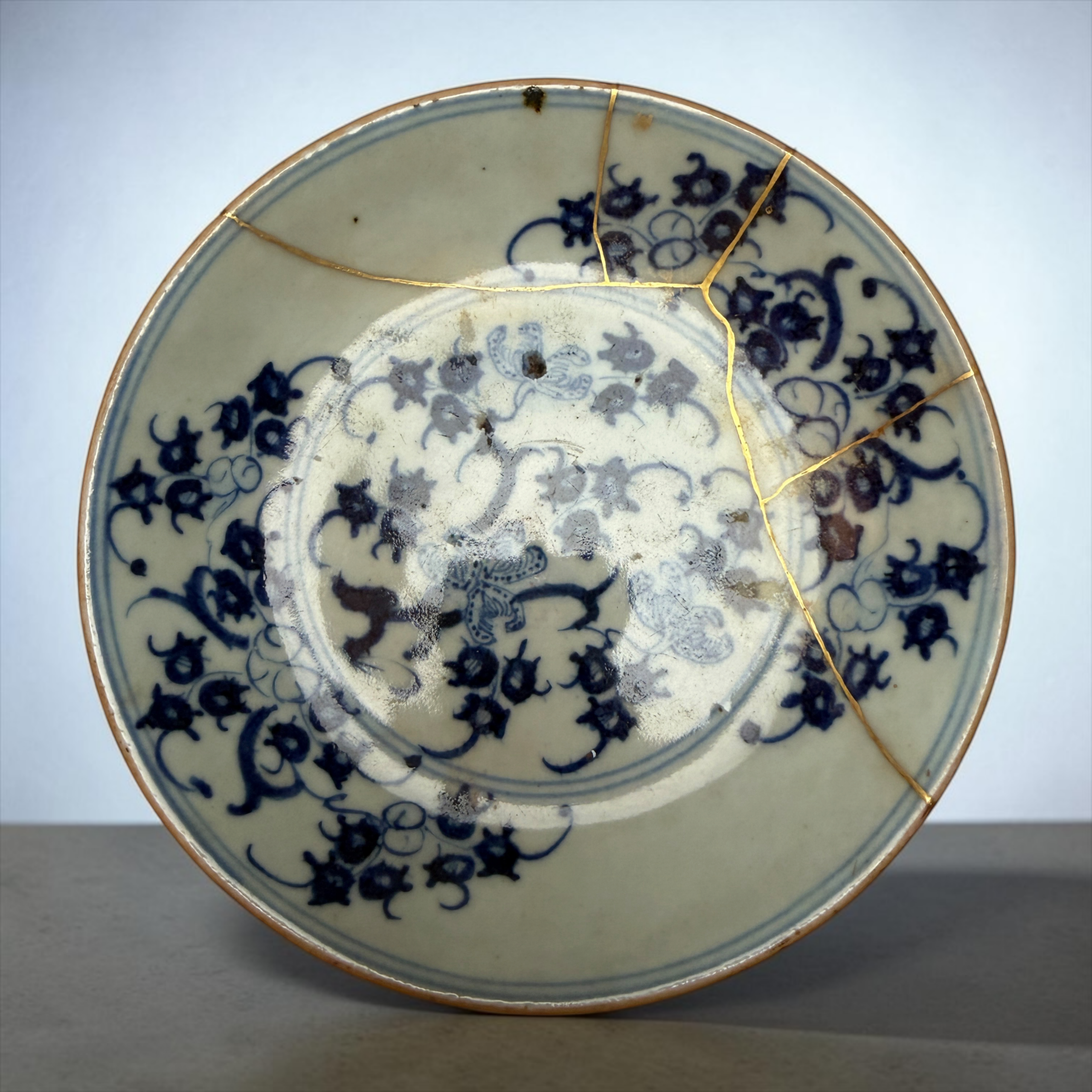 IMG_2561 | Kintsugi by Myriam GREFF Decorative blue-and-white porcelain plate with gold crack repair (kintsugi), shown from the edge against a gray background